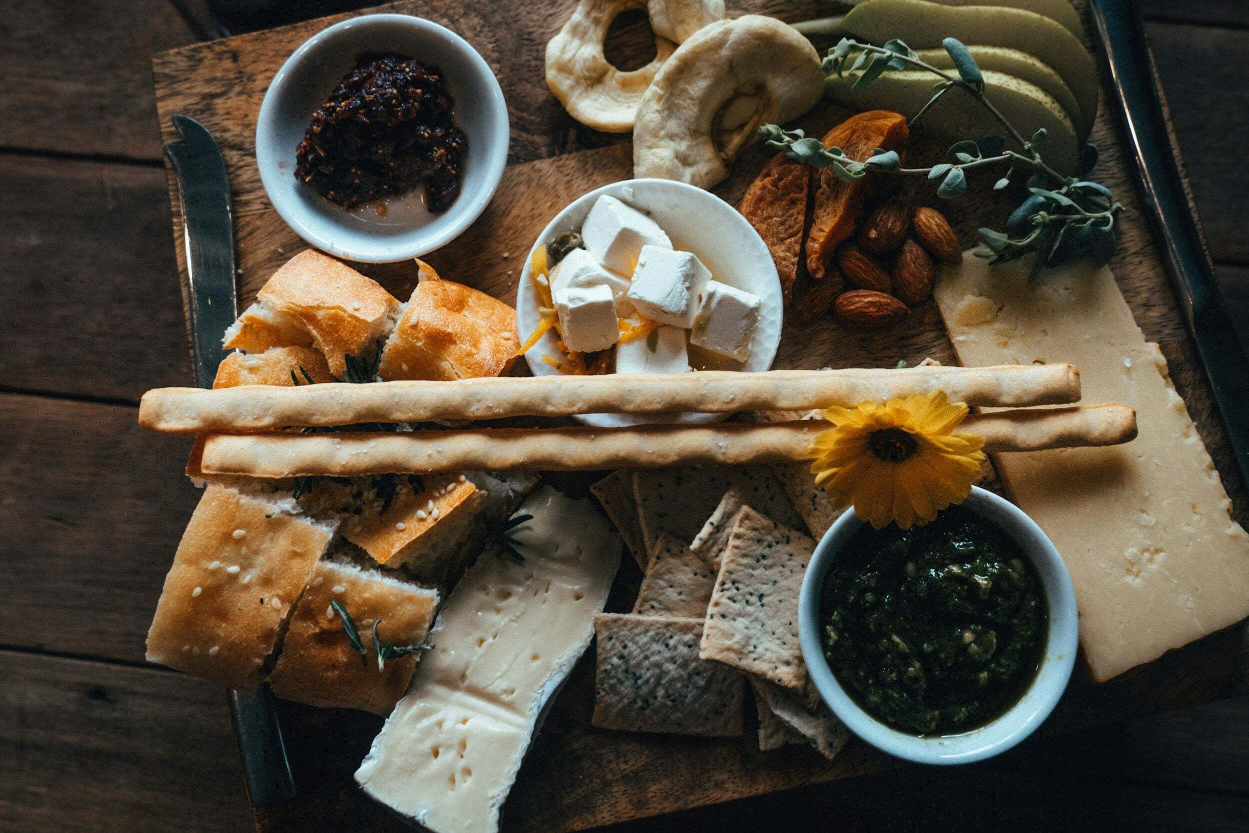 platou de lemn cu branzeturi de diferite texture, paine si dulceata langa flori galbene si crengute cu frunze verzi